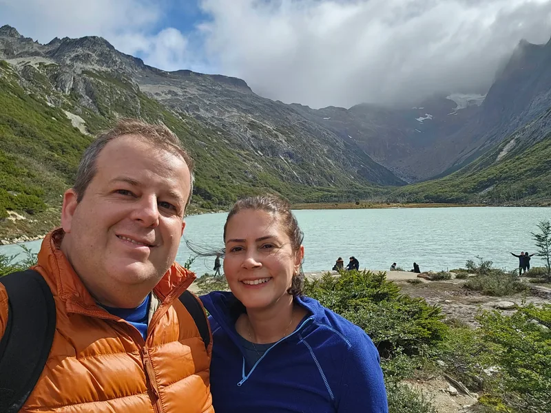 Charles e Denise em selfie diante do lago, com algumas pessoas distantes na margem.