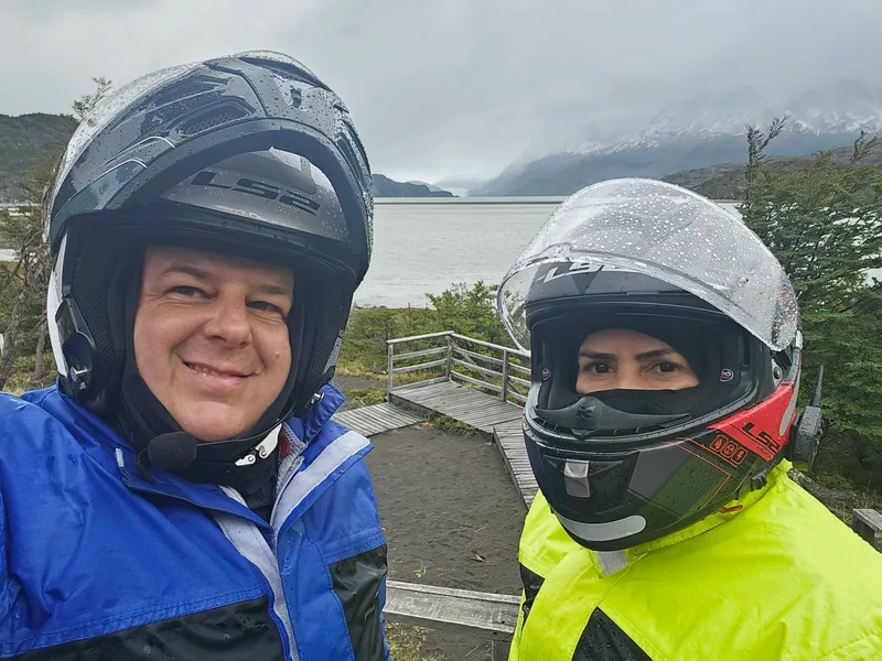 Charles e Denise de capacete, com paisagem de lago e montanhas ao fundo.