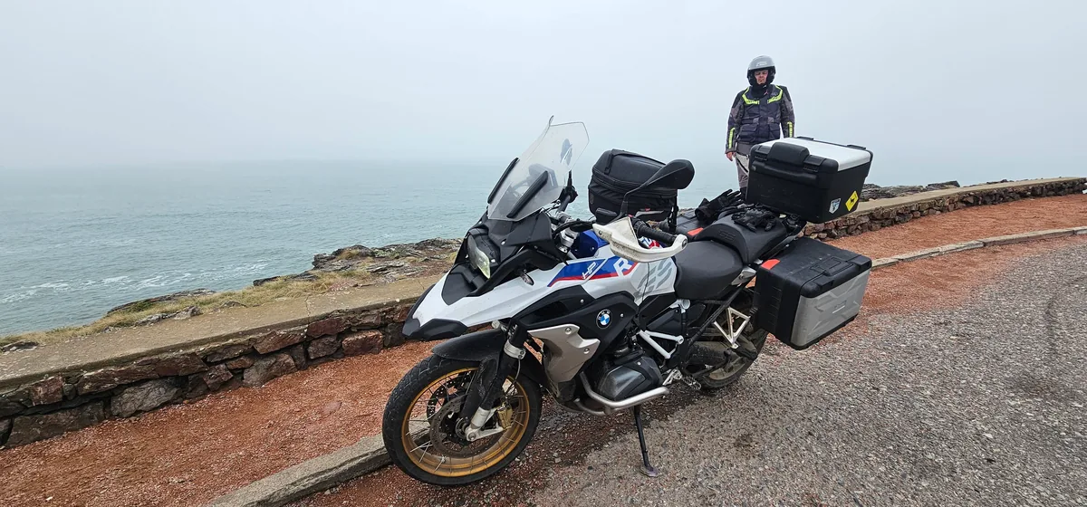Moto BMW estacionada em mirante à beira-mar, com um motociclista ao fundo.