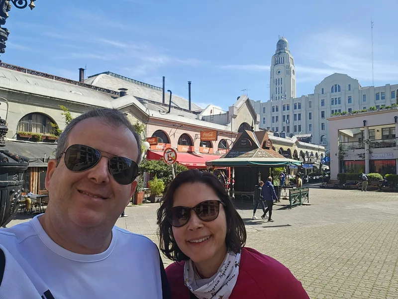 Casal em selfie na praça do Mercado del Puerto, com prédios históricos ao fundo.