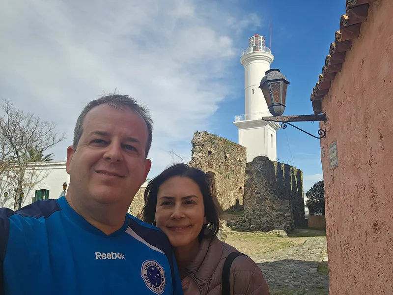 Casal em selfie diante de um farol branco e de ruínas de pedra, em Colônia del Sacramento.
