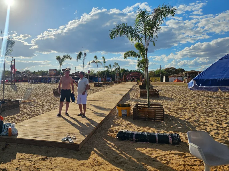 Márcio e Chiquinho em passarela de madeira, com mesas e guarda-sóis da praia ao fundo.